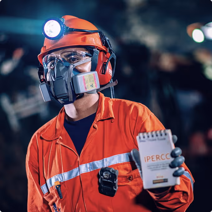 Worker wearing an orange protective suit, helmet with light, safety goggles, and gas mask holding a notepad labeled IPERCC.