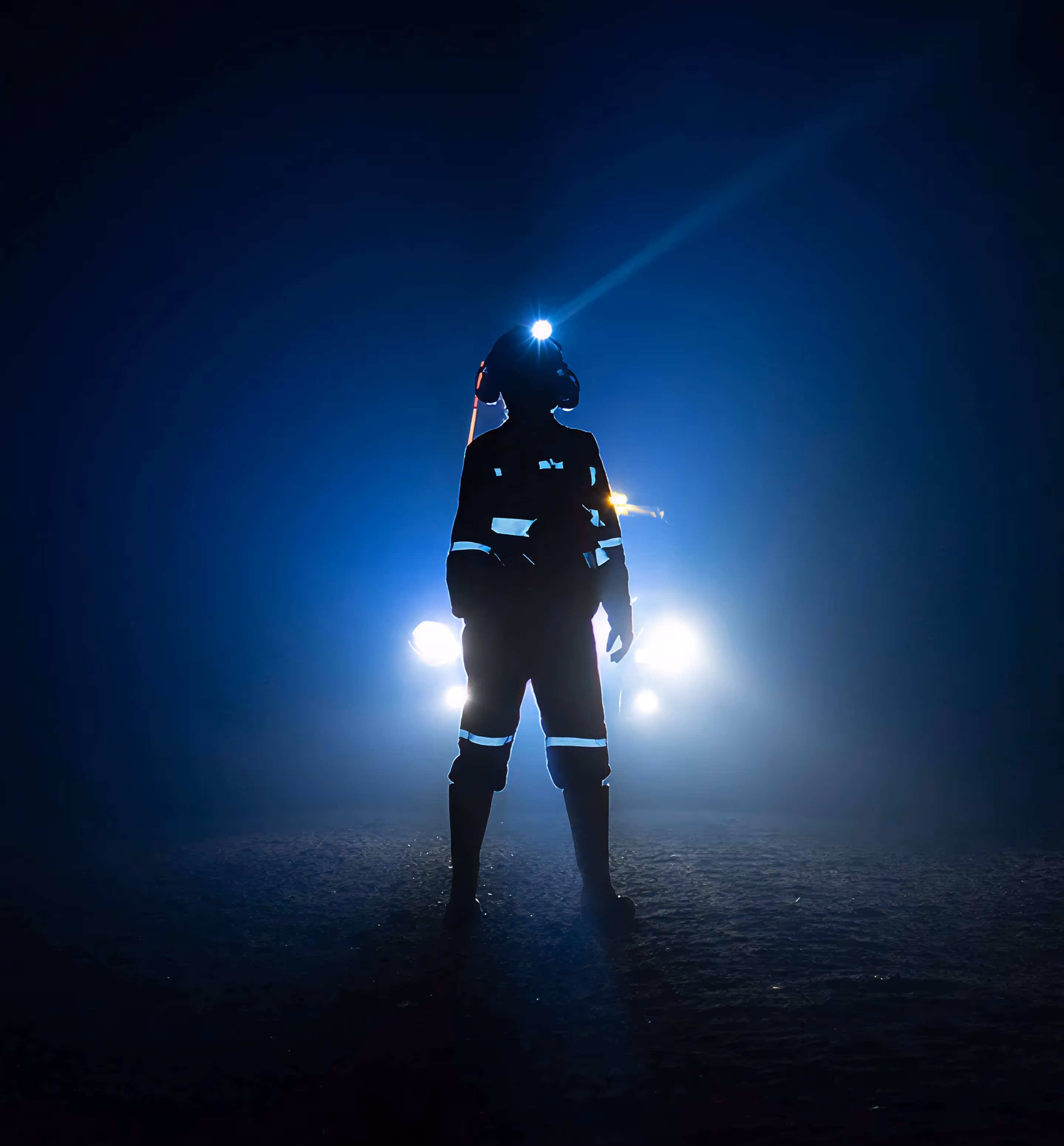 Silhouette of a person wearing a helmet with a headlamp, standing in front of bright headlights in a dark, foggy environment.