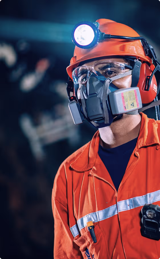 Worker wearing orange safety gear, respirator mask, safety glasses, and a helmet with a headlamp in a dimly lit environment.