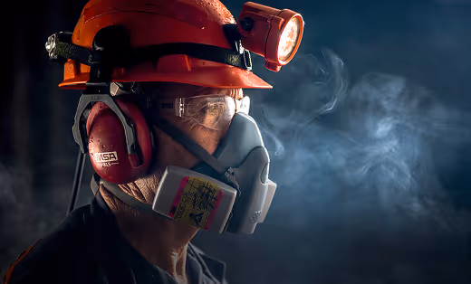 Worker wearing an orange hard hat with a headlamp, protective goggles, a respirator mask, and ear protection, with smoke in the background.