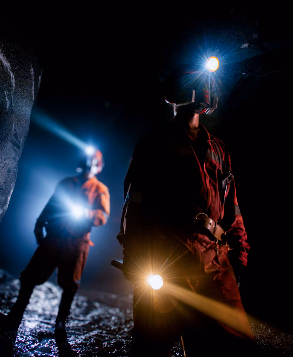 Two miners wearing helmets with headlamps and holding flashlights inside a dark underground mine.
