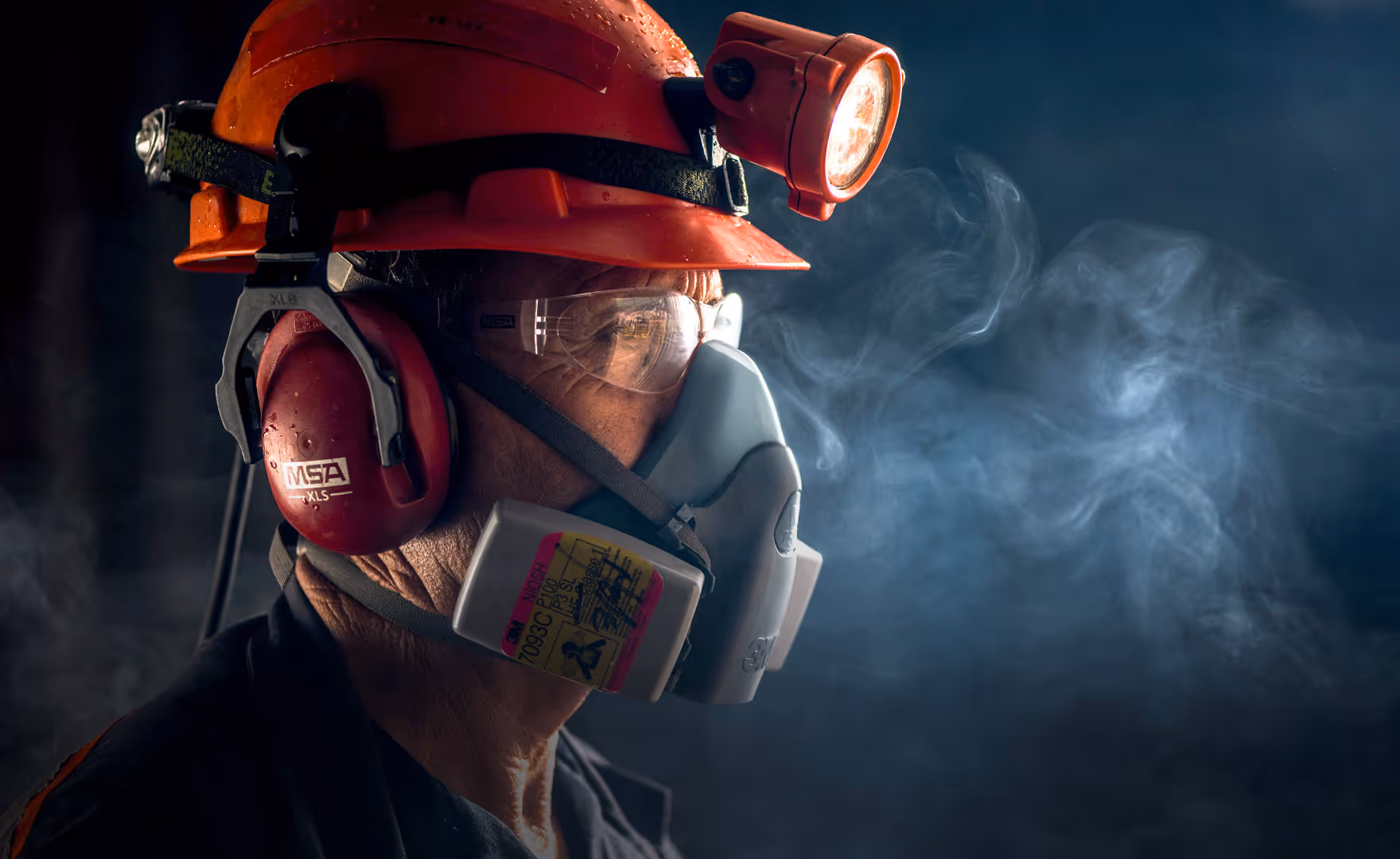 Worker wearing an orange helmet with a headlamp, red ear protection, safety glasses, and a protective respirator mask with smoke visible in the background.