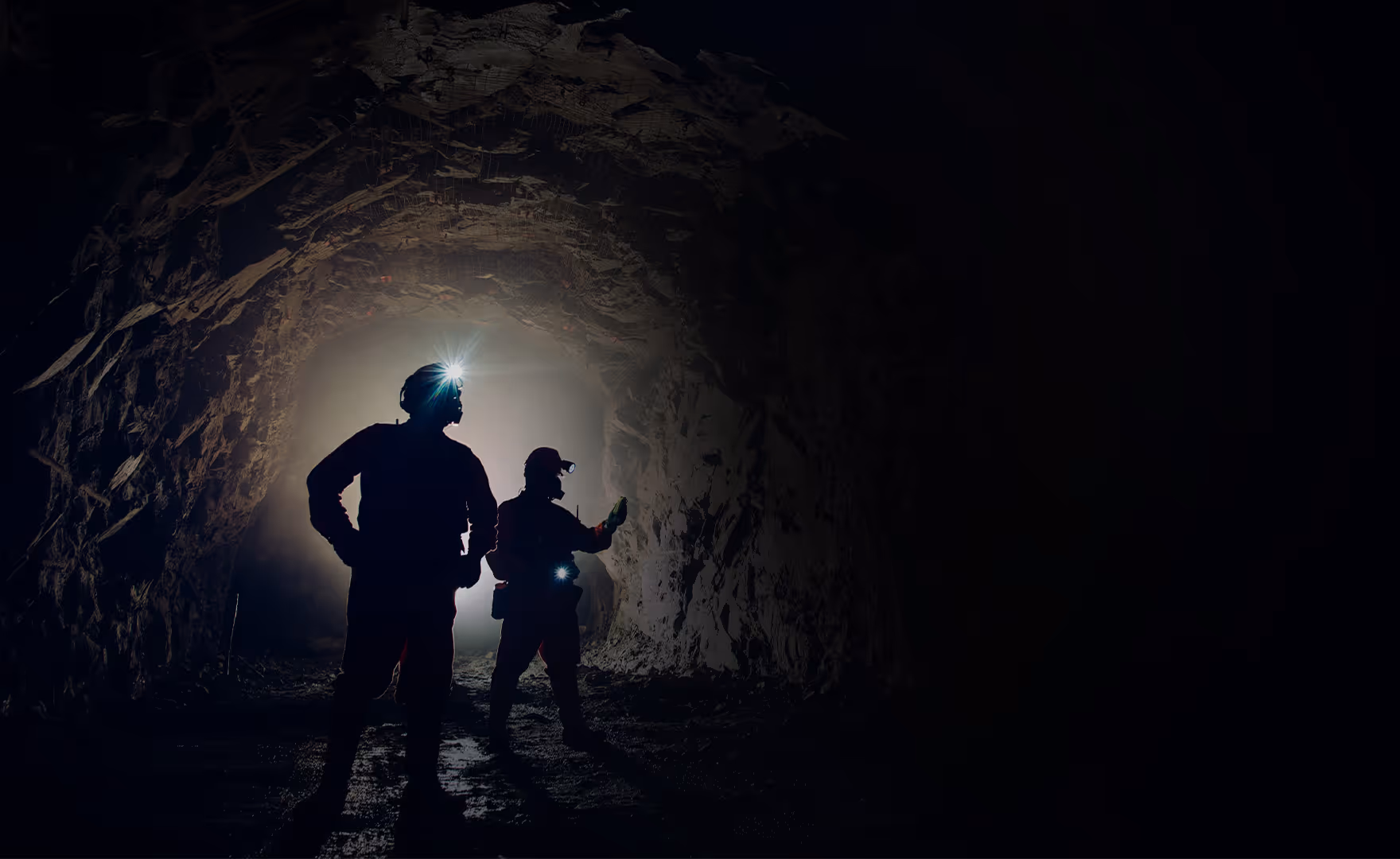 Two miners wearing helmets with headlamps standing inside a dimly lit rock tunnel.