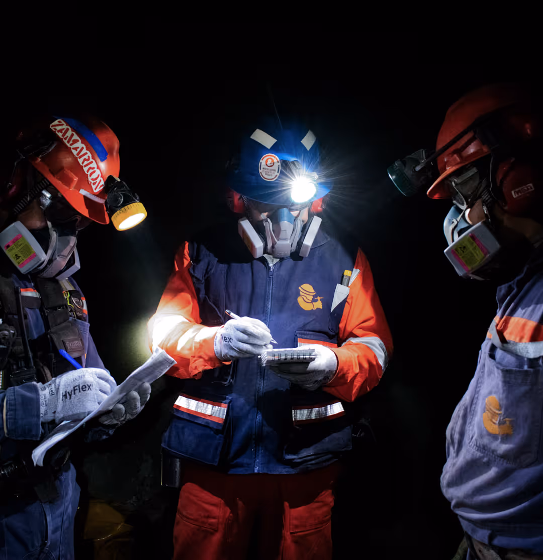 Three workers wearing protective gear and helmets with headlamps writing notes in a dark environment.