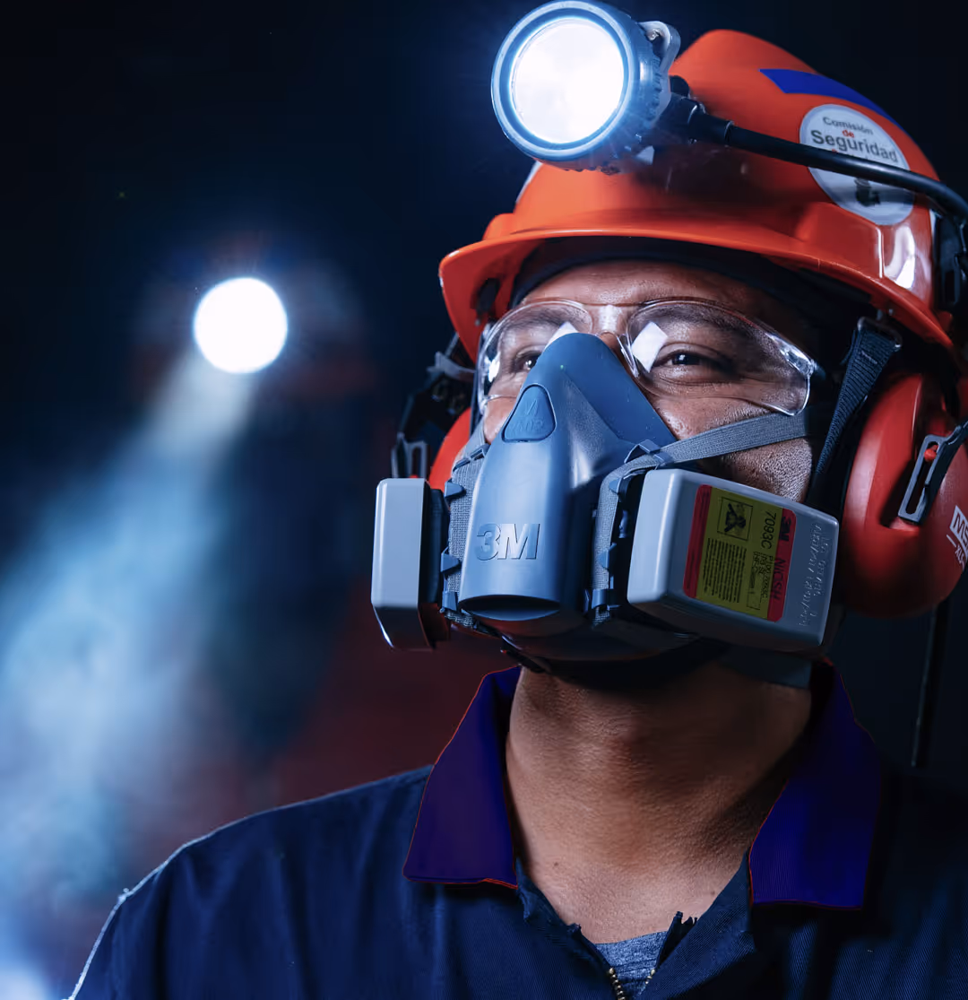 Worker wearing a red safety helmet with a headlamp, protective goggles, a respirator mask, and ear protection against a dark background.