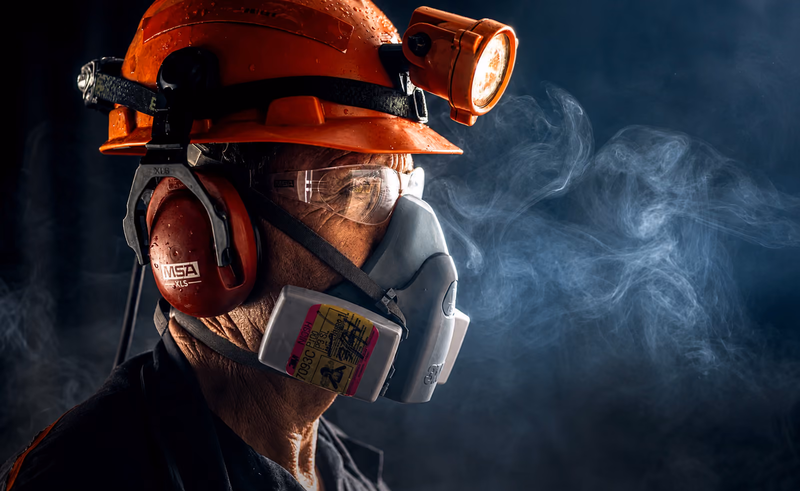 Worker wearing a red helmet, safety goggles, earmuffs, and a respirator mask in smoky environment.