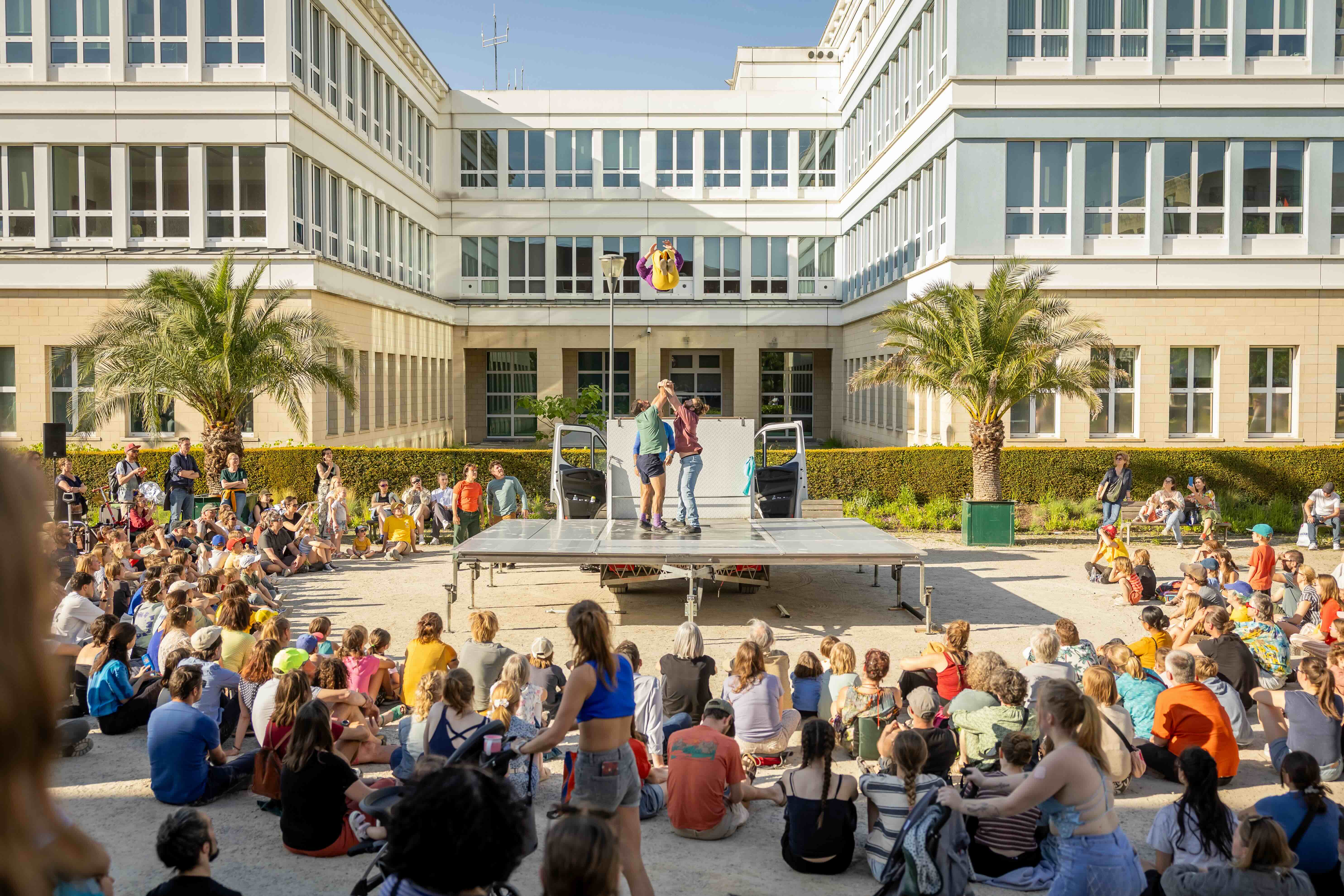 Een plein vol kinderen die kijken naar een circusvoorstelling, waarbij iemand van de artiesten in de lucht geworpen wordt.