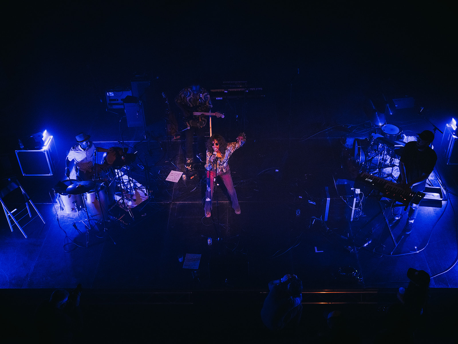 Een zangeres met zonnebril zingt in een microfoon op podium, de muzikanten rond haar zijn amper verlicht - zij staat in een spotlight.