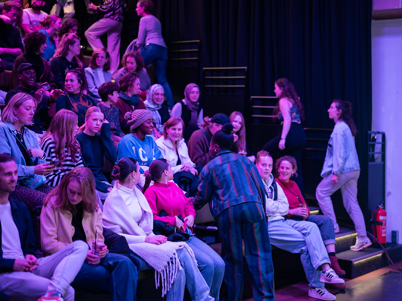Een auditorium gevuld met mensen die op oplopende banken zitten onder paars licht. De aanwezigen in casual kleding zijn in gesprek, wat een levendige sfeer creëert.