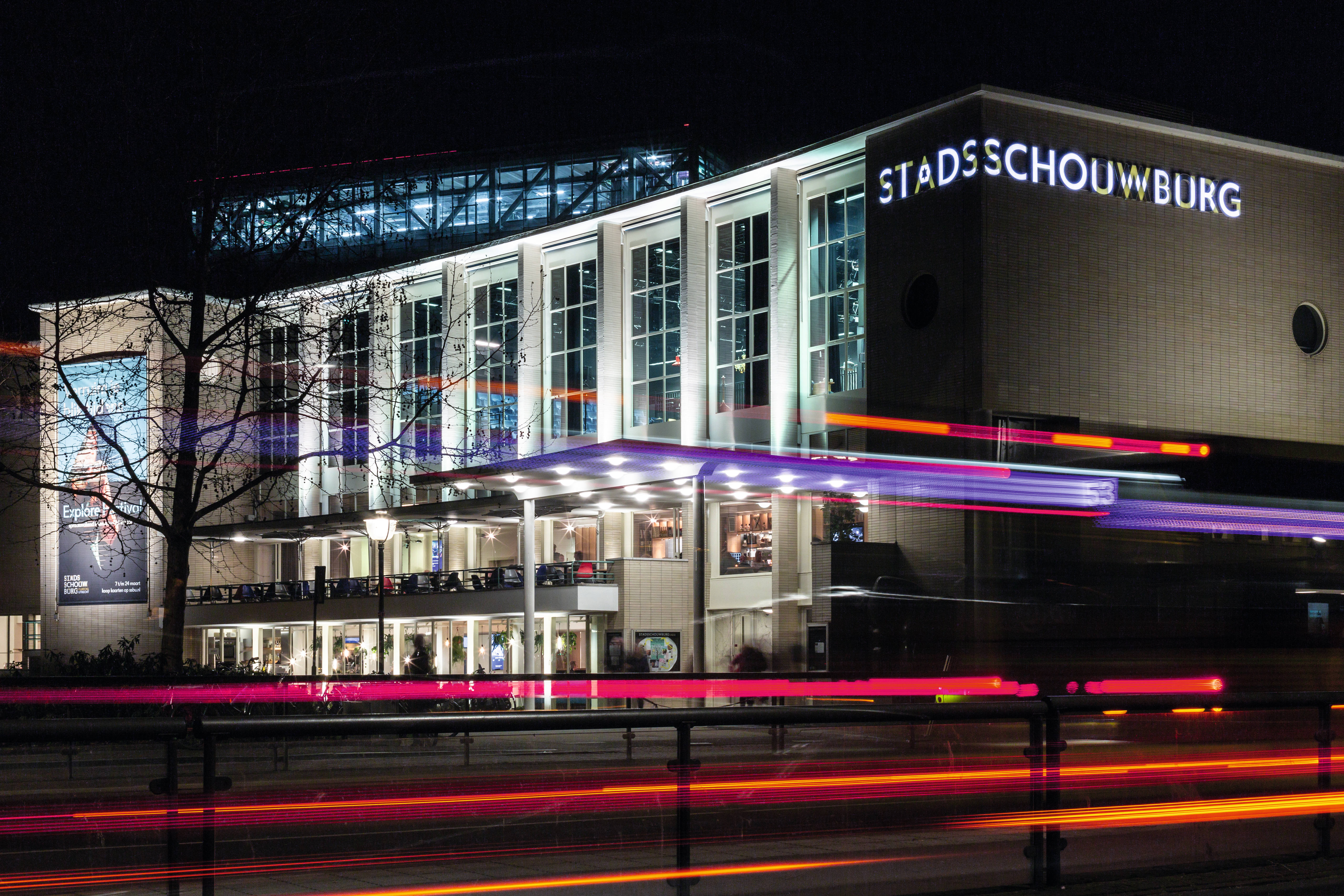 Stadsschouwburg Utrecht verlicht in de avond, met lichtsporen van voorbijrijdend verkeer en zicht op de moderne glazen gevel.