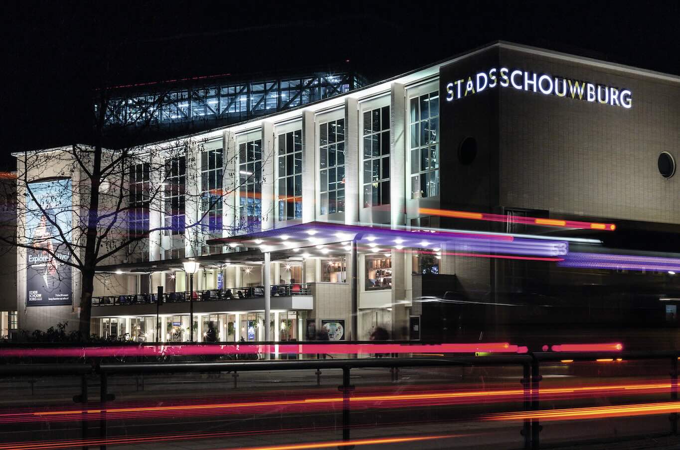 Stadsschouwburg Utrecht verlicht in de avond, met lichtsporen van voorbijrijdend verkeer en zicht op de moderne glazen gevel.