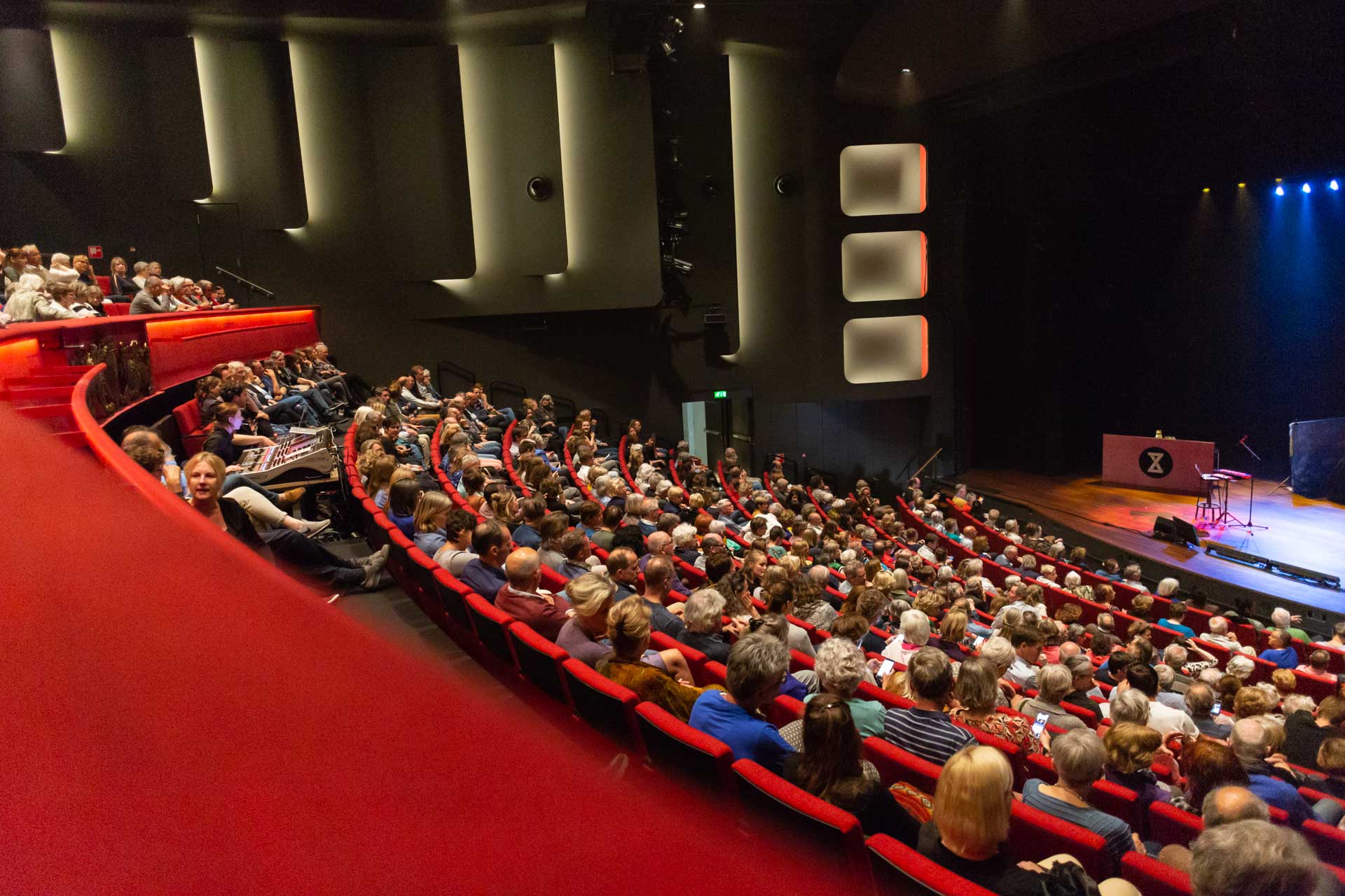 Publiek in de Grote Zaal van Stadsschouwburg Utrecht tijdens een voorstelling, met uitzicht op het podium en de rode theaterstoelen.