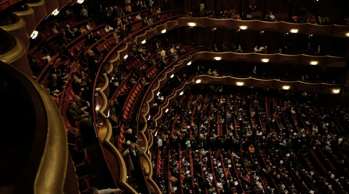 Audience gathered in a large classical theater with multiple balconies and red seating, just before a performance begins.