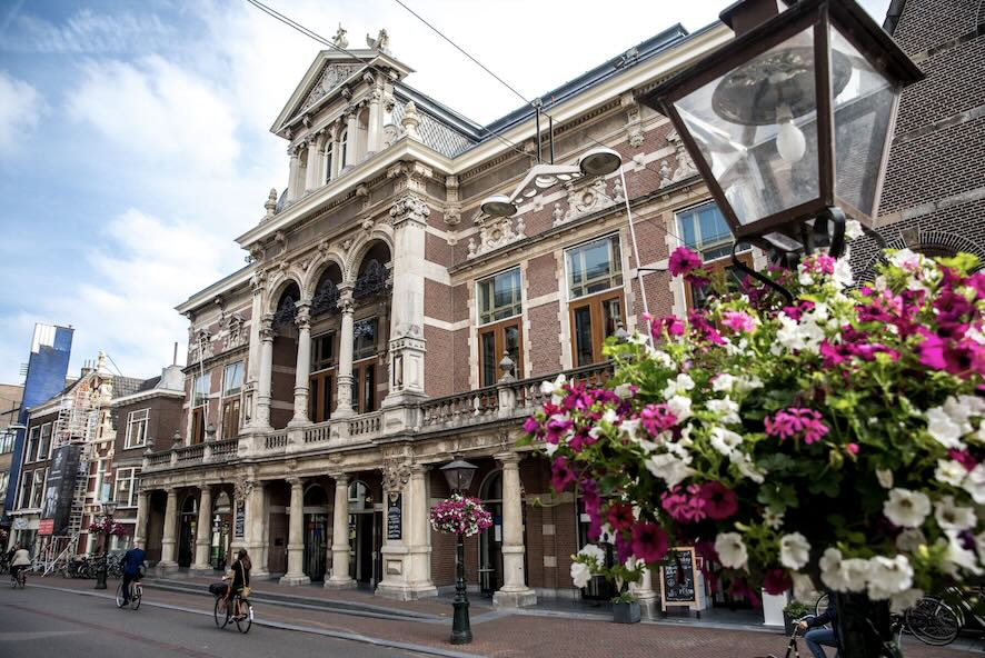 De voorgevel van de Leidse Schouwburg – Stadsgehoorzaal aan een levendige straat, met fietsers en kleurrijke bloemen op de voorgrond.