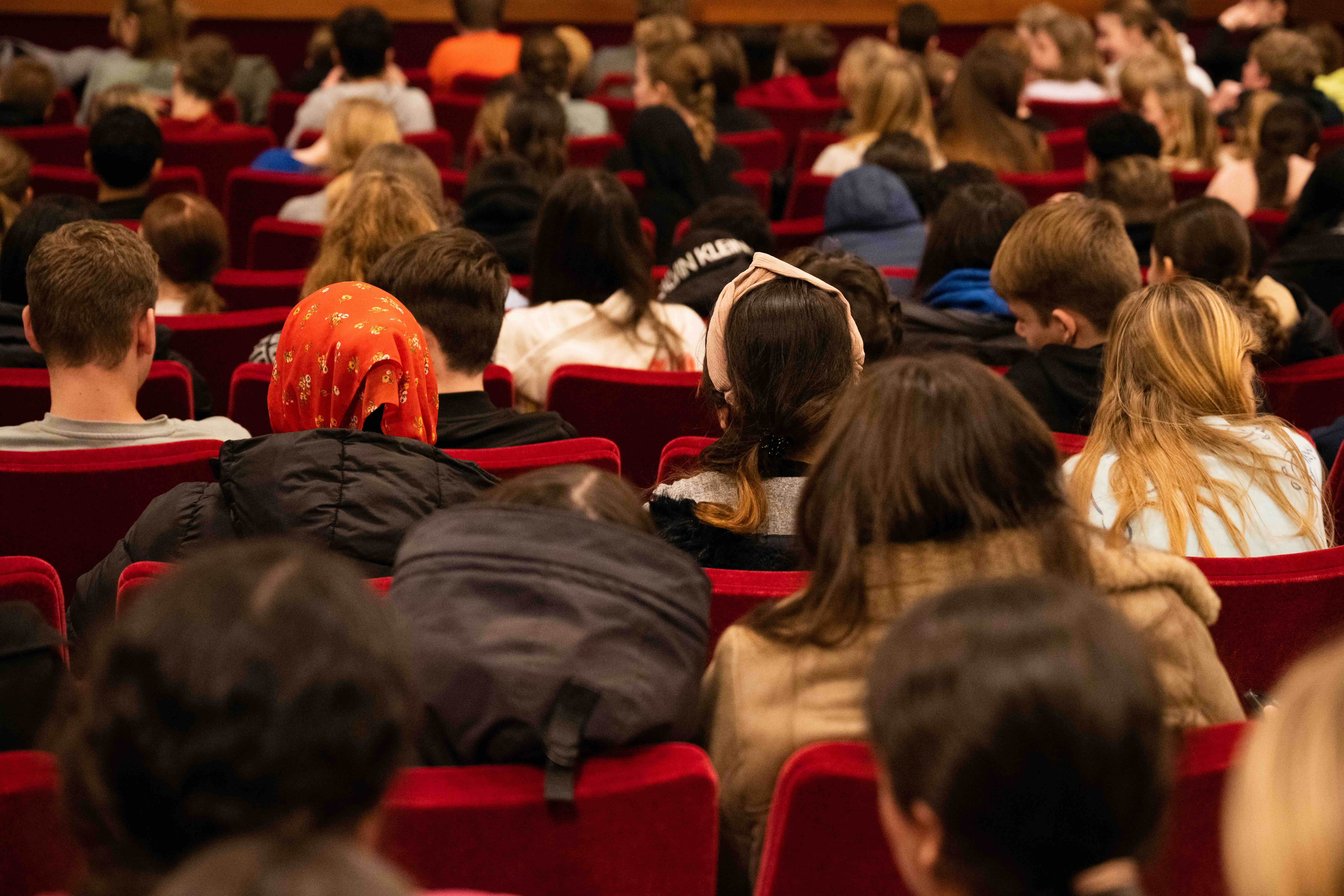 Een diverse groep toeschouwers, vooral jongeren, zit dicht op elkaar in rode theaterstoelen tijdens een voorstelling of evenement.