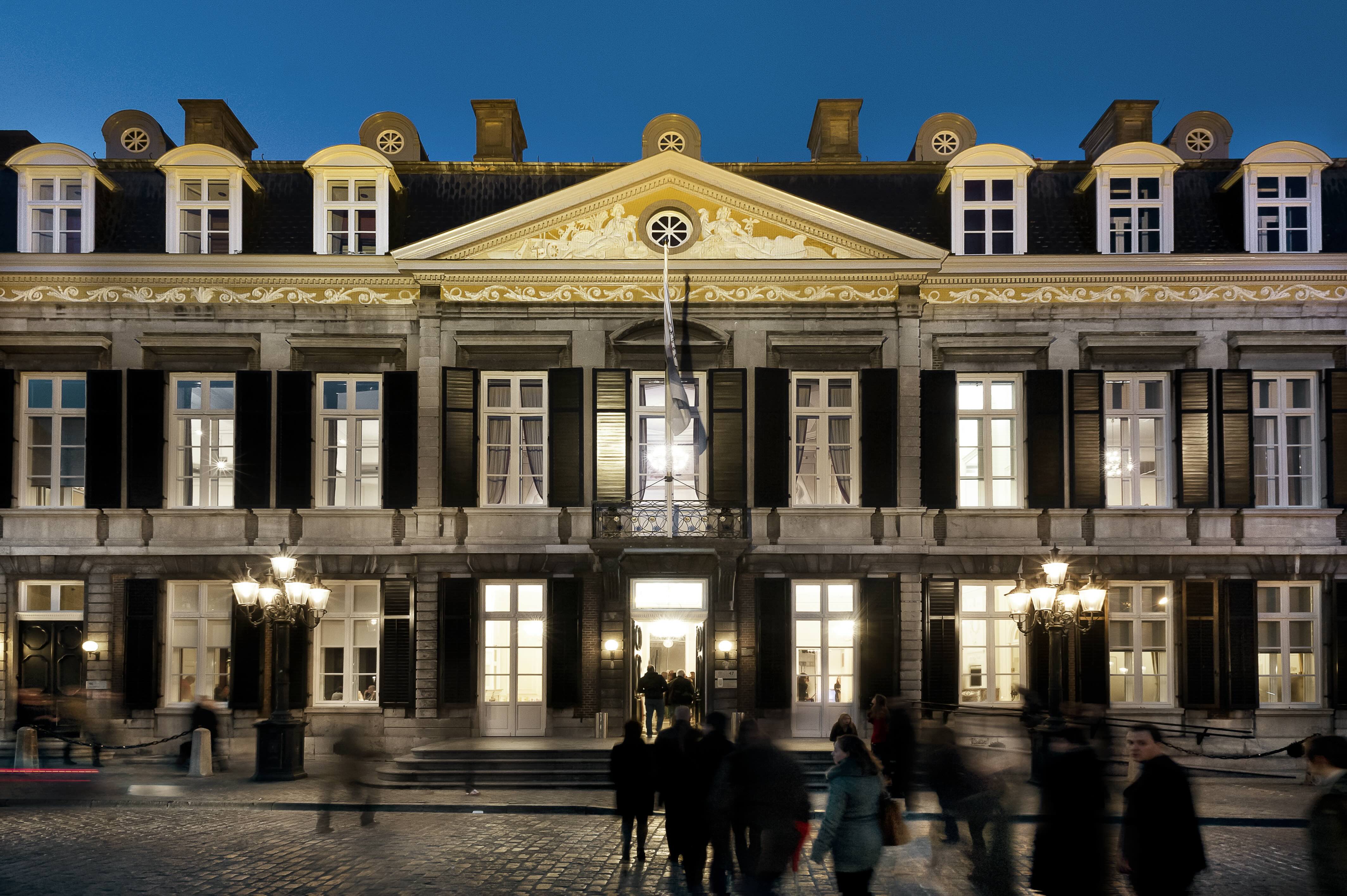 The illuminated façade of Theater aan het Vrijthof in Maastricht at dusk, with people walking across the square in front of the building.