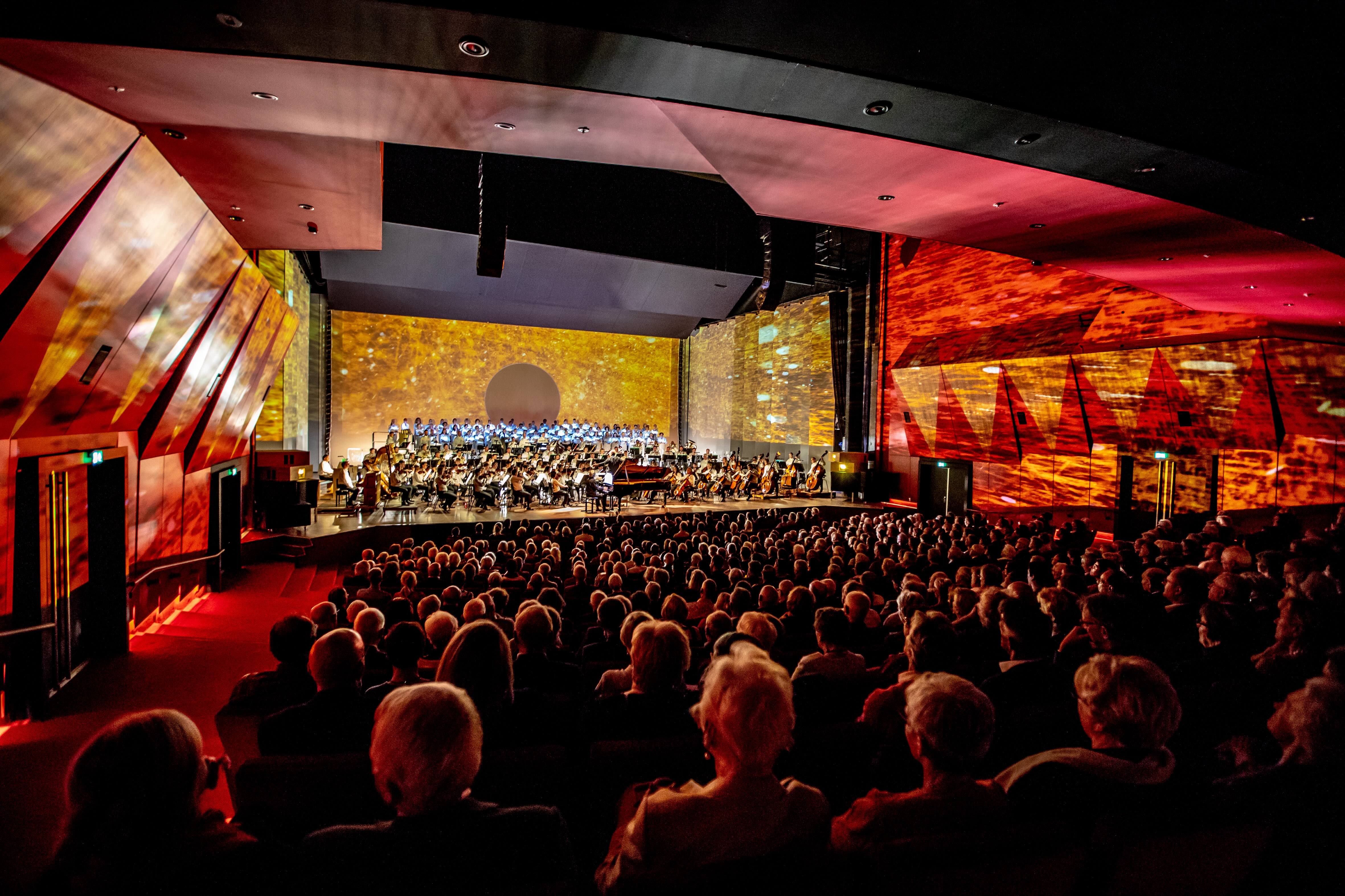The Papyruszaal at Theater aan het Vrijthof filled with audience members, watching a large orchestra and choir perform amid colorful projections on the walls.