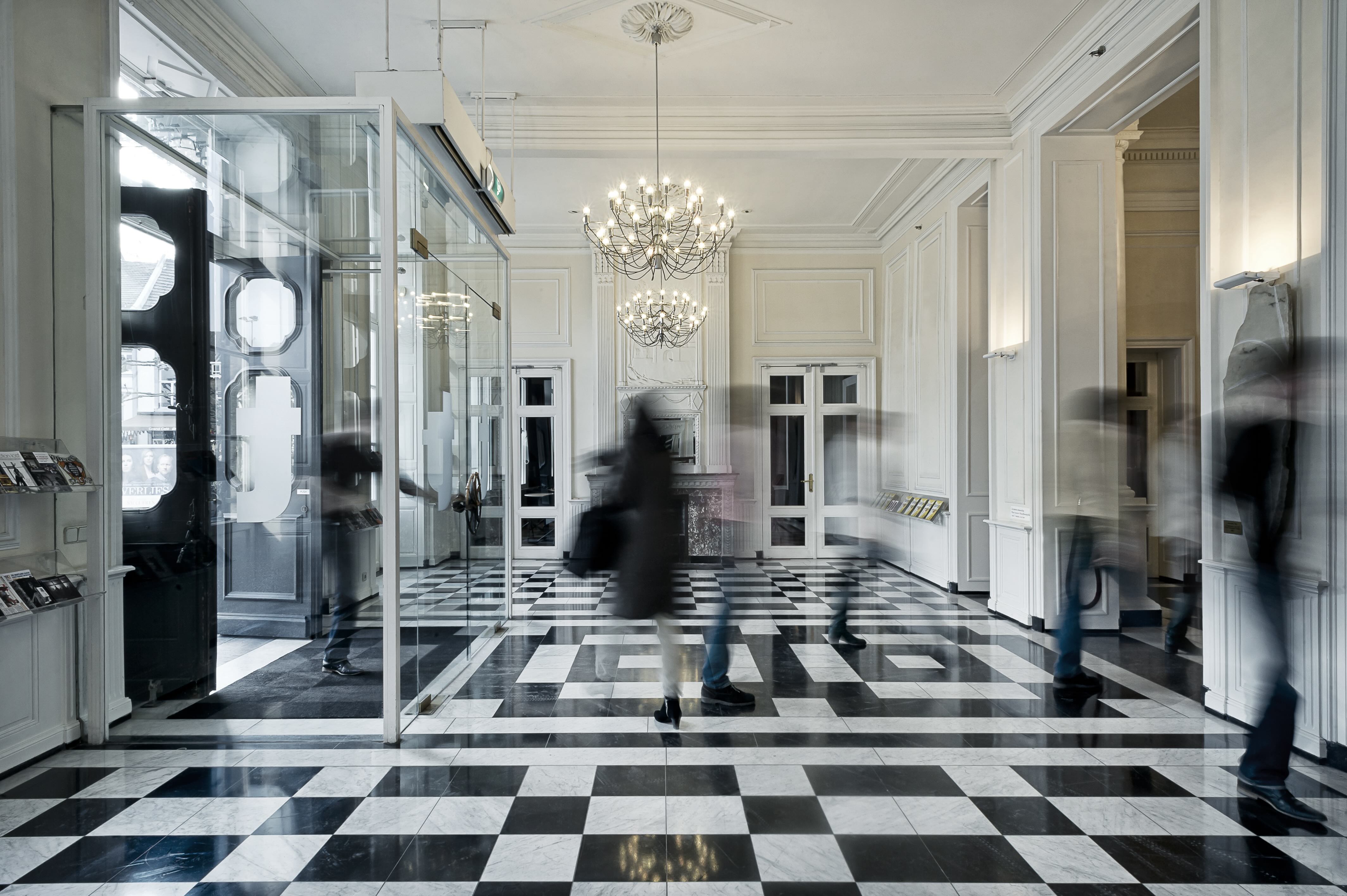 The bright, classically styled foyer of Theater aan het Vrijthof with a black-and-white marble floor, chandeliers, and visitors moving through the space.