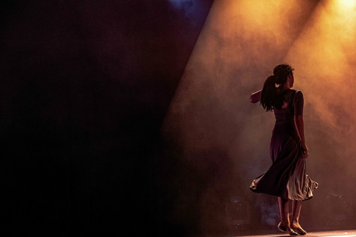A dancer on stage, seen from behind, moving in warm orange and purple stage lighting against a dark background.