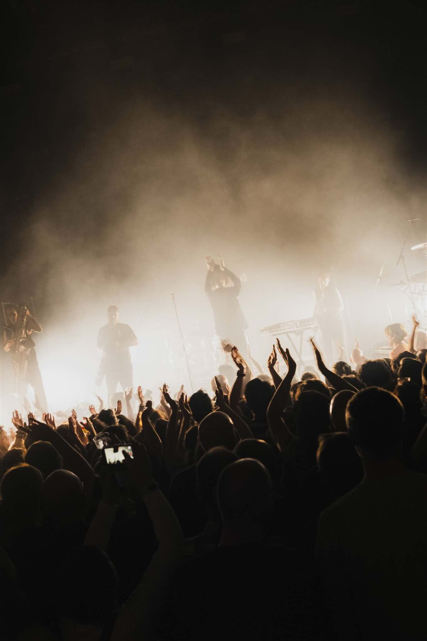 Public les bras levés lors d'un concert au Depot, avec des artistes sur scène enveloppés de fumée et de lumière.