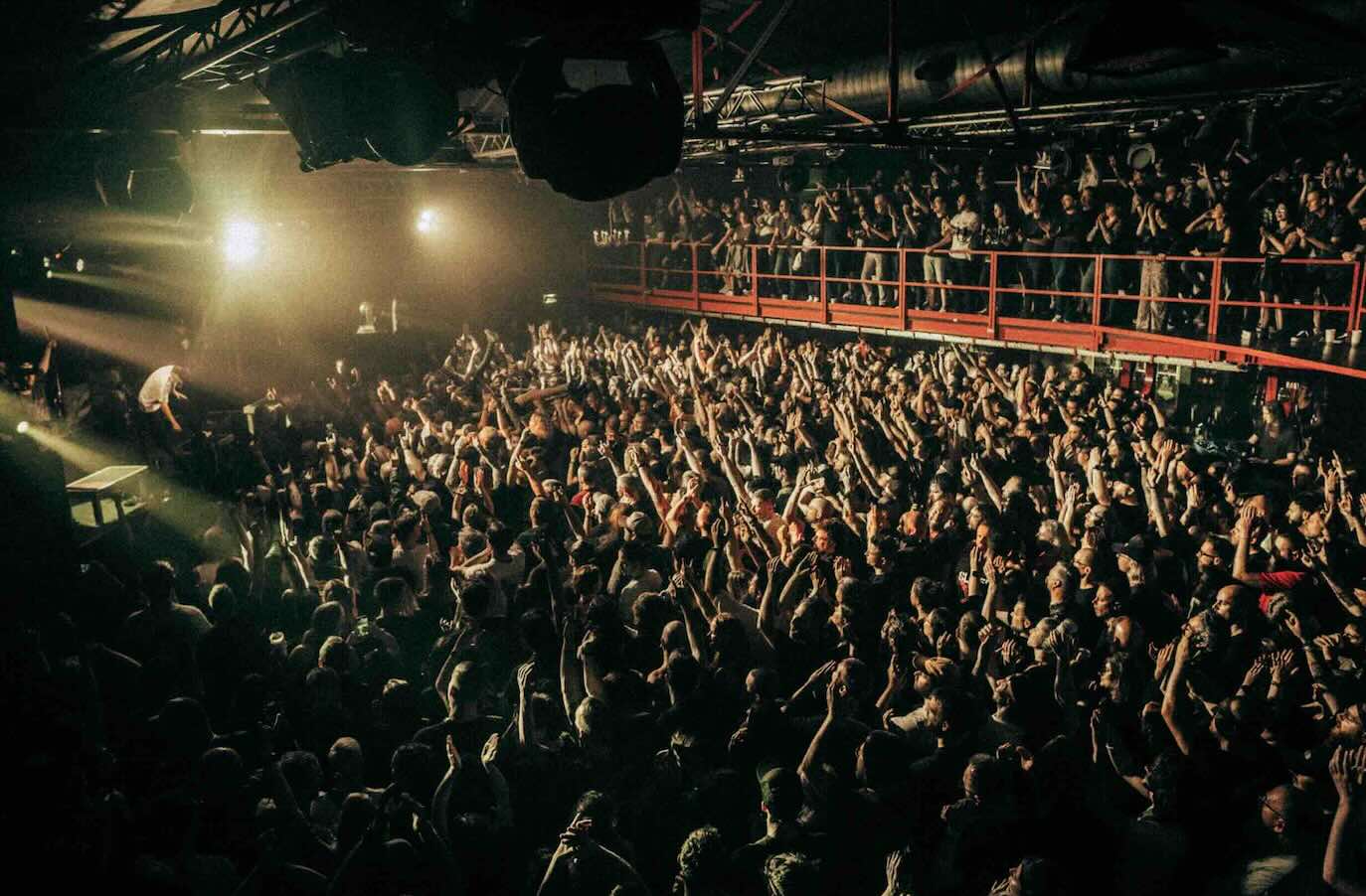 A sold-out concert venue with an energetic crowd raising their hands, viewed from the floor with a packed balcony above.