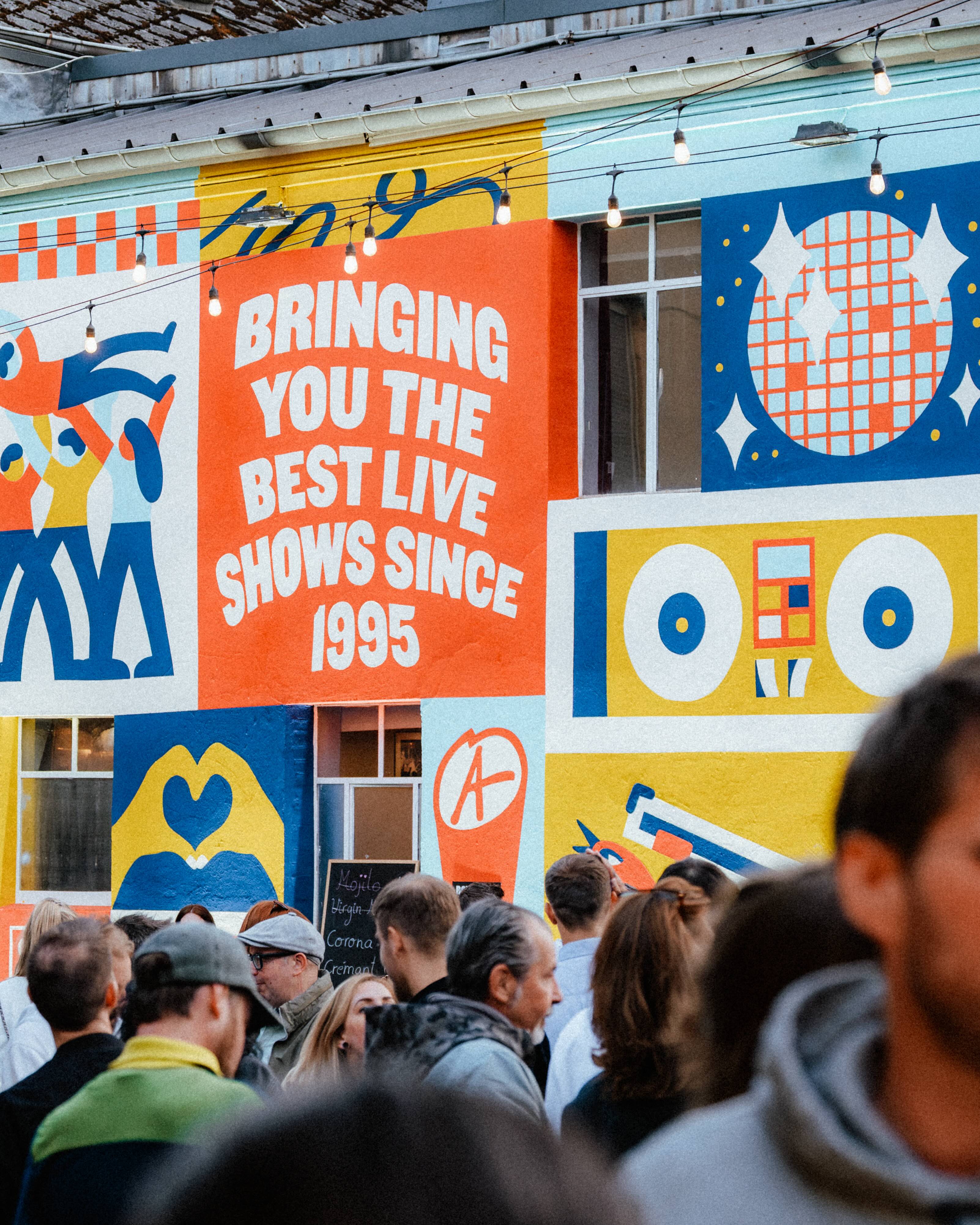 Visitors gather in front of a colorful mural reading “Bringing you the best live shows since 1995”.