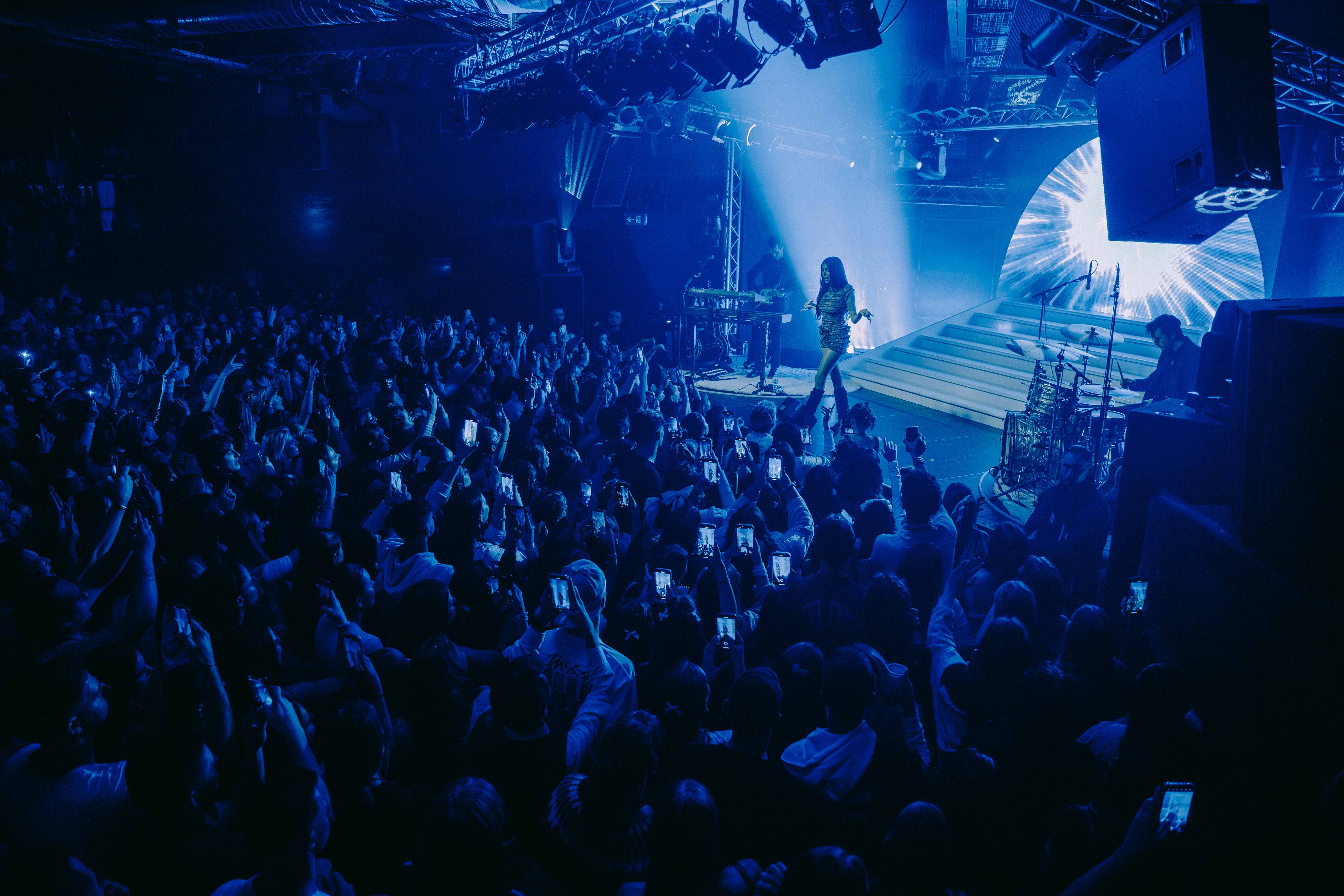 Artist performing under blue stage lights in front of a packed crowd recording the show on their smartphones.