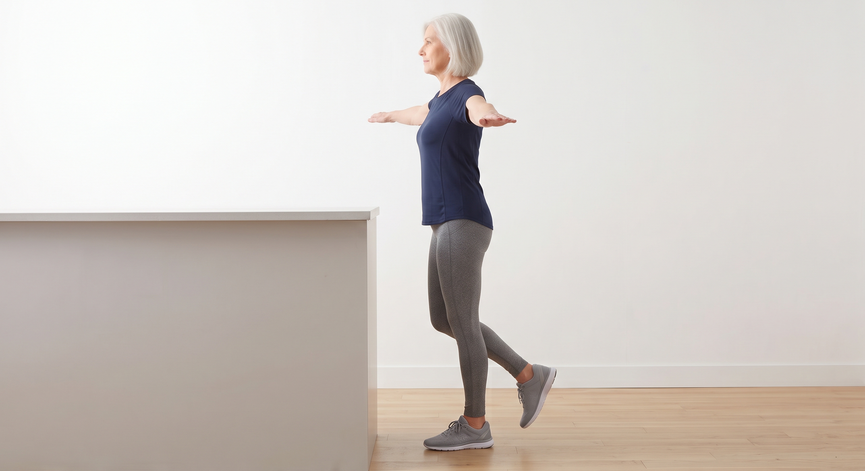 Older woman balancing on one leg a step away from a kitchen counter with both arms extended out to the sides, performing the hands-free progression of single leg standing balance