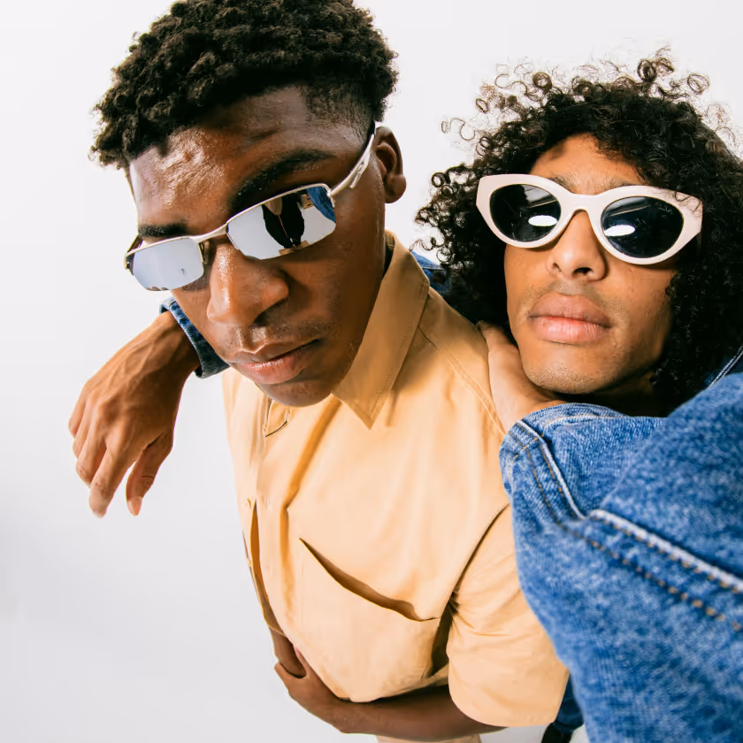Two young men wearing sunglasses, one in a beige shirt and the other in a denim jacket posing together.