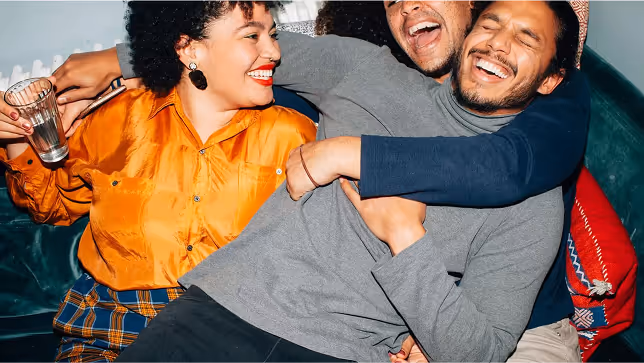 Three friends sitting closely on a couch, laughing and hugging each other joyfully.