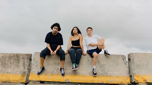 Three young people sitting on a large concrete barrier under a cloudy sky.