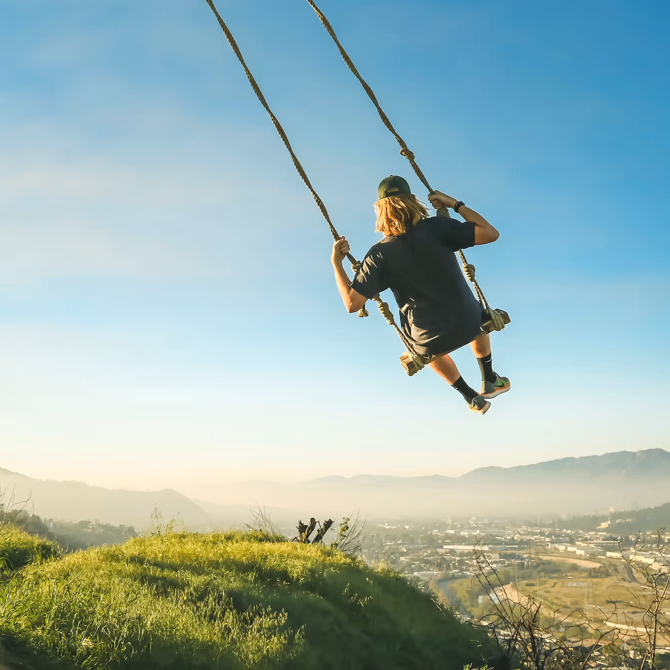 Person swinging on a large rope swing over a grassy hill with a city and mountains in the background under a clear blue sky.