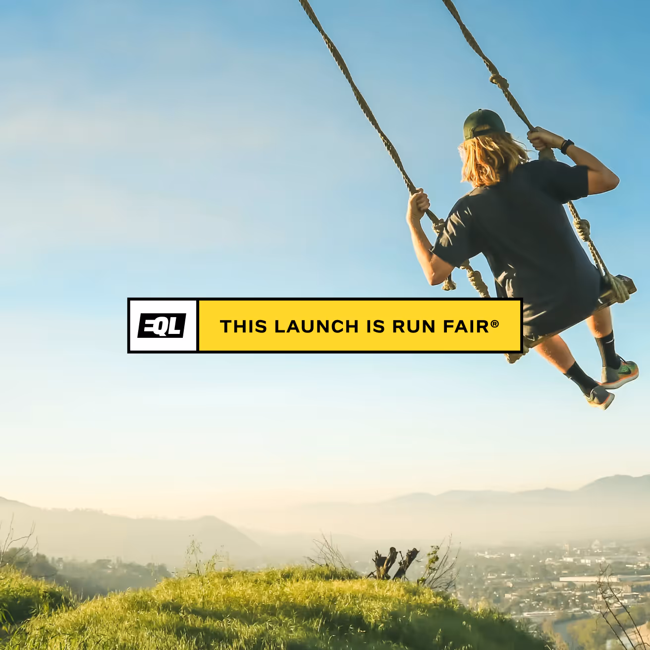 Person with long blond hair wearing a green cap swinging on a rope swing above green hills with a city and mountains in the distance under a clear sky.