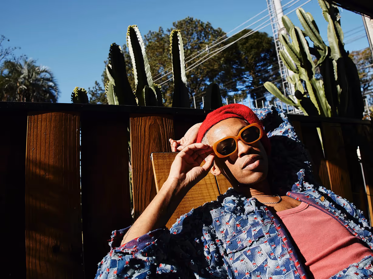 Person in a red beanie and orange sunglasses reclining outdoors against a wooden fence with tall cacti in the background.