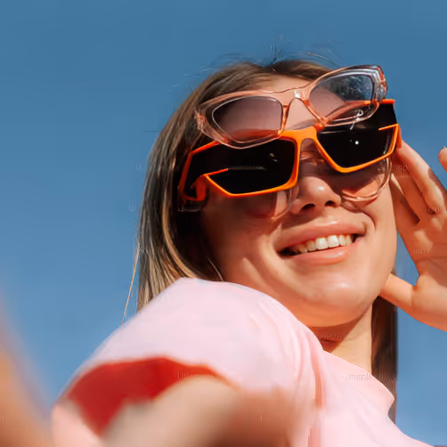 Woman smiling wearing two pairs of sunglasses stacked on her face against a blue sky background.