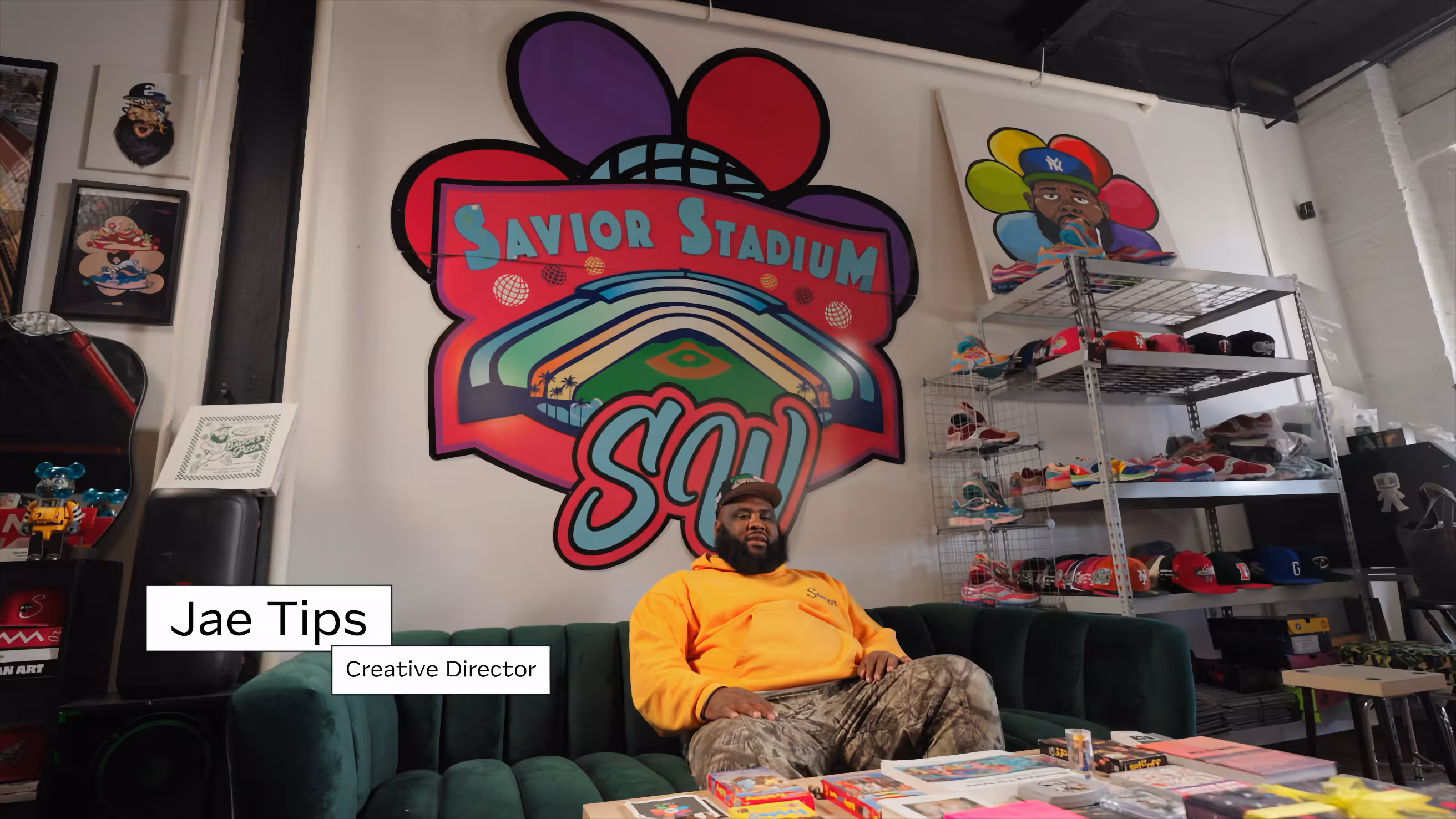 Man wearing an orange hoodie and camouflage pants sitting on a green velvet couch in front of a colorful 'Savior Stadium SOU' wall mural, surrounded by sneaker shelves and artwork.