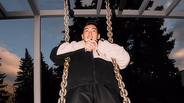 Man sitting on a chain swing outdoors against a dark evening sky, looking upwards with hands clasped.