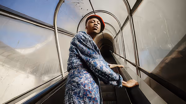 Man wearing a red beret and patterned coat standing on an escalator in a tunnel-like glass enclosure.