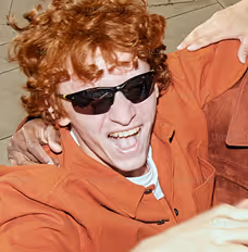 Person with curly red hair wearing black sunglasses and an orange jacket, smiling with mouth open.