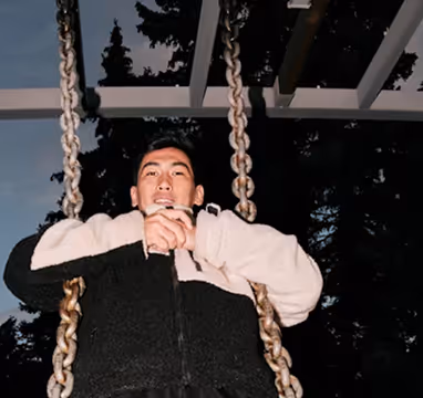 Man wearing a black and white fleece jacket sitting on a swing with metal chains against a dark background.