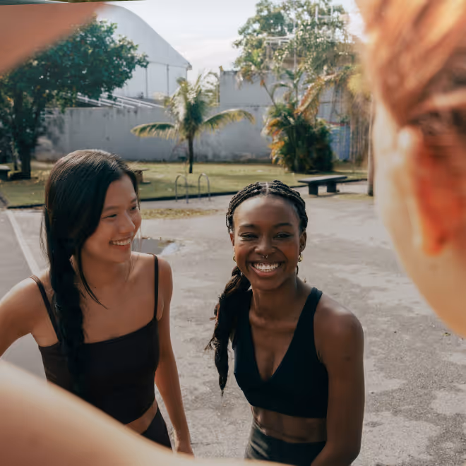 Two young women smiling and standing outdoors in casual athletic wear with greenery and structures in the background.