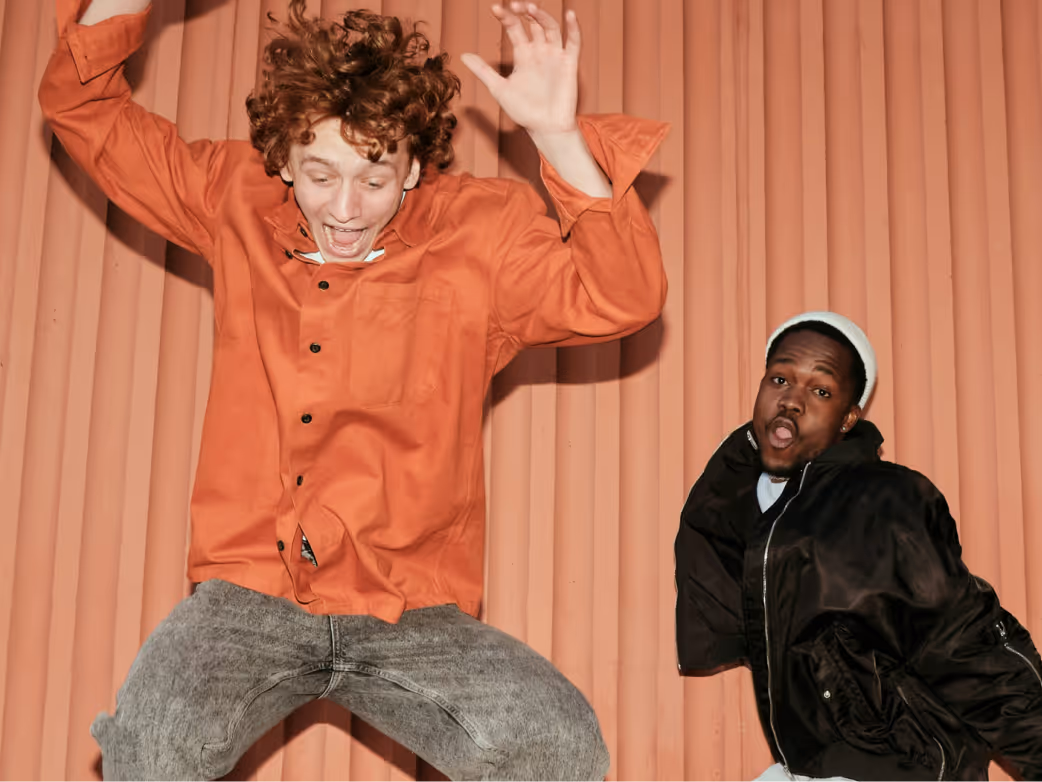 Two young men expressing surprise and excitement, one jumping wearing an orange shirt, the other reacting wearing a black jacket and white beanie.