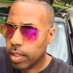 Man wearing reflective pink sunglasses and a black shirt standing near a car outdoors.