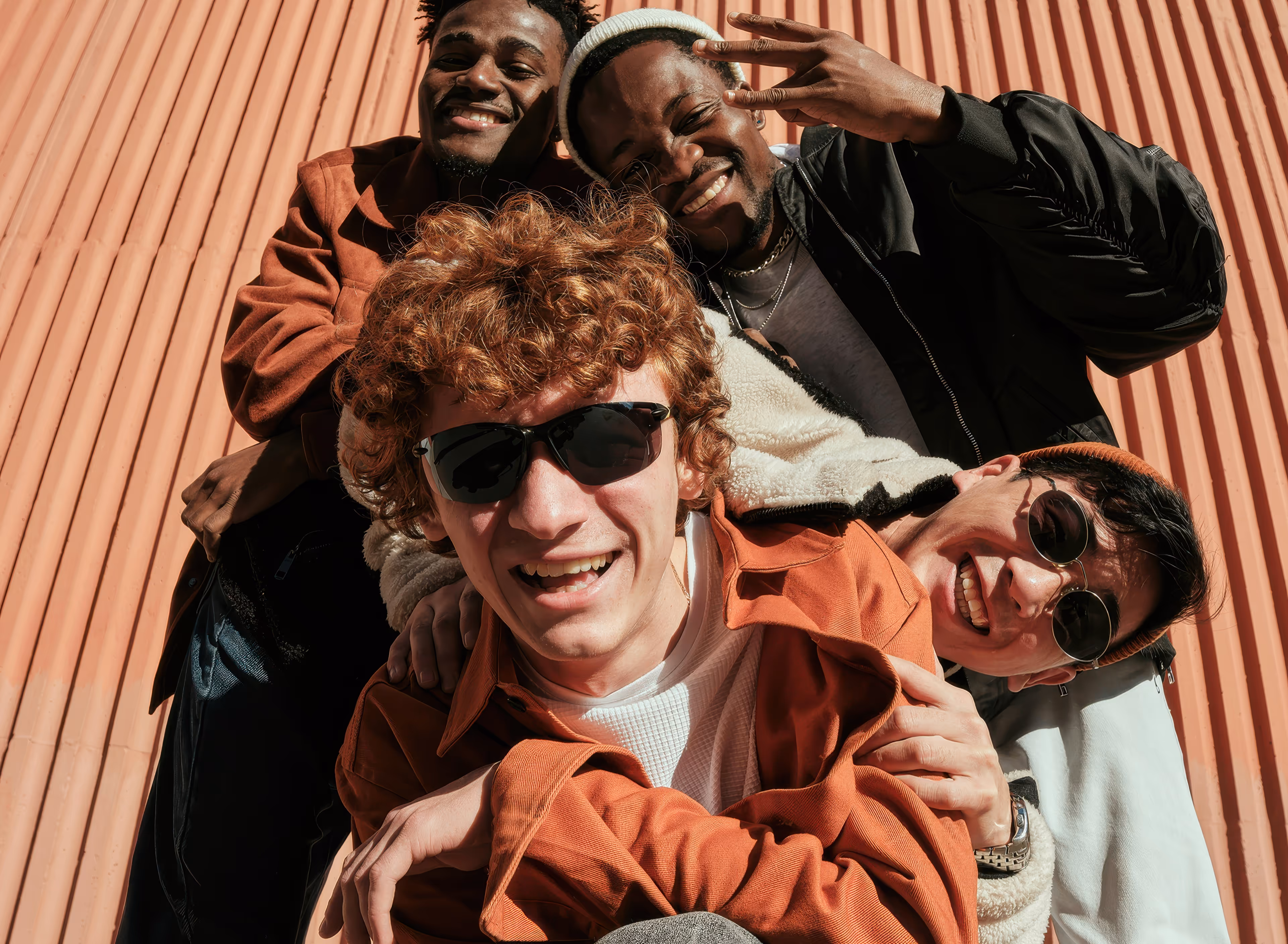 Four young men smiling and posing closely together against a corrugated orange wall.