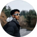 Man wearing white sunglasses and dark jacket outdoors with blurred trees and rocks in background.