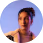 Woman with dark hair in a loose bun against a purple-blue background.