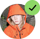 Person wearing an orange hooded jacket with a serious expression in front of greenery.