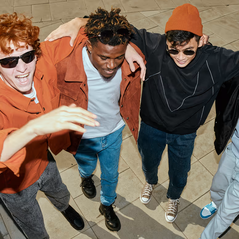 Four young men with arms around shoulders walking outdoors on tiled ground, smiling and wearing sunglasses.