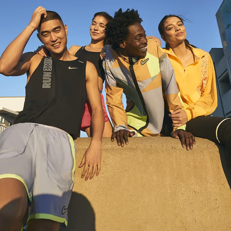 Four young adults smiling outdoors, dressed in sporty Nike apparel, leaning on a concrete ledge.