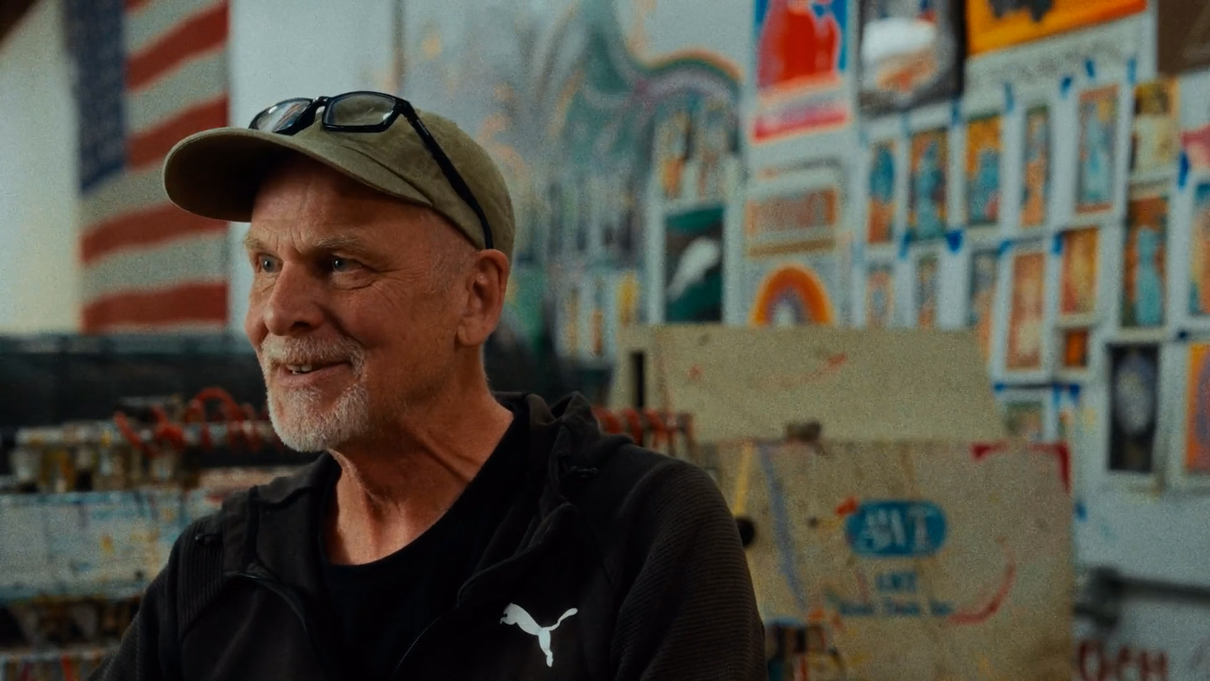 Smiling older man with white beard and cap wearing a black Puma jacket in an art-filled room.